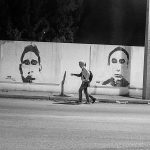 2 young men walk along the wall with graffiti of 2 unknown men faces